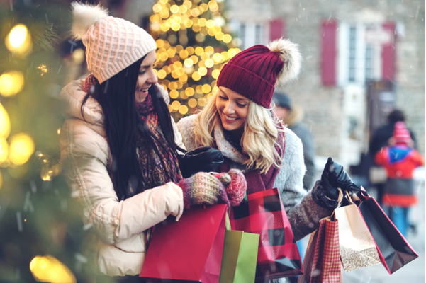 Deux femmes debout à l'extérieur d'une petite entreprise en hiver, tenant des sacs de courses et souriant. Ils profitent d’une journée de shopping local.