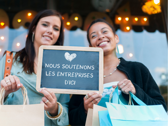 Deux femmes souriantes et tenant des sacs de courses se tiennent devant une entreprise canadienne locale tenant un tableau noir sur lequel est écrit « Nous soutenons les entreprises d'ici ».