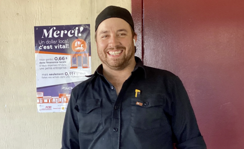 Le propriétaire d'Attaché-Remorque, membre de la FCEI au Québec, debout à côté de l'affiche d'impact local de la FCEI dans son commerce et souriant.